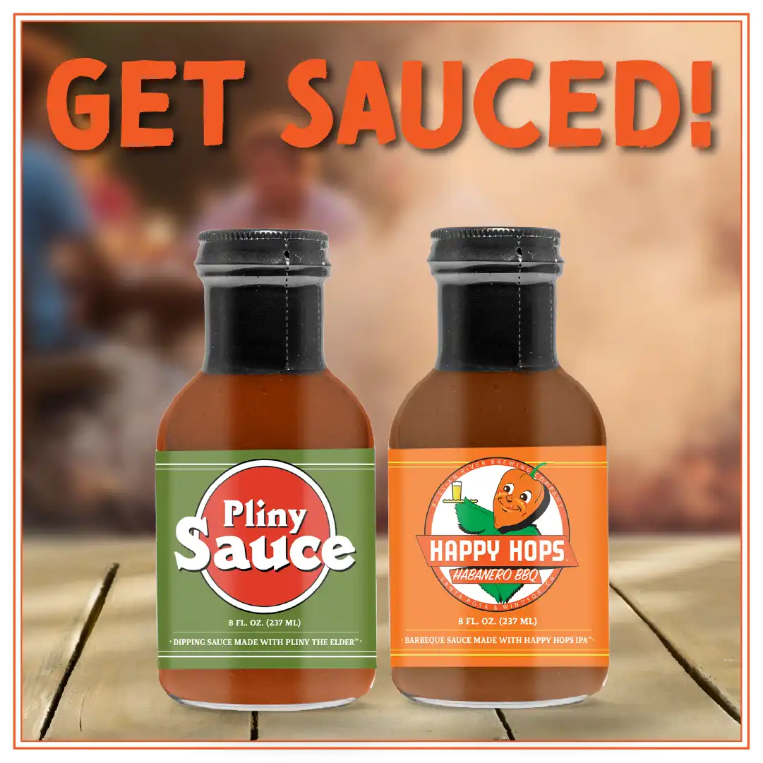 A picture of Pliny Sauce and Happy Hops Habanero BBQ Sauces in bottles on a wood table with a group of people barbequing in the background.